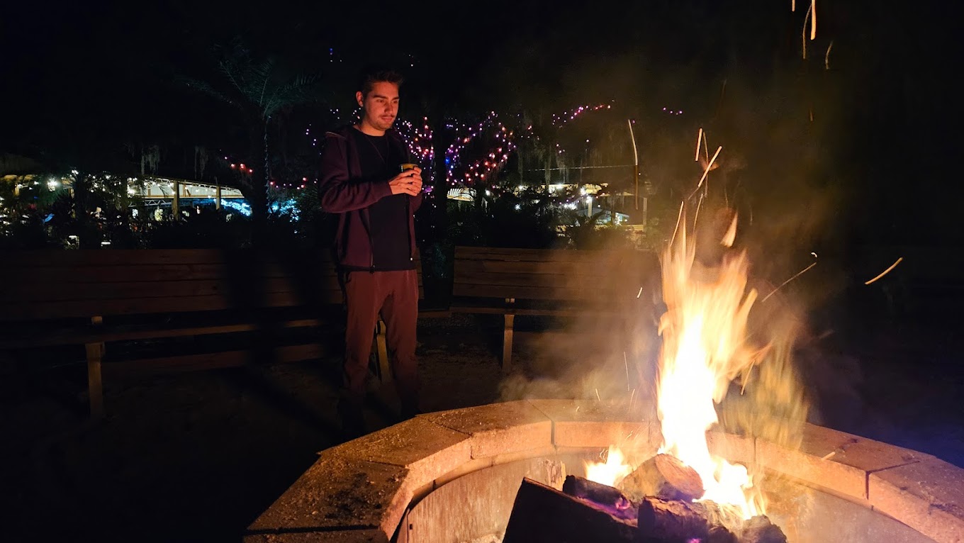 A young man by a fire pit at night holding a cup, warm evening camp atmosphere