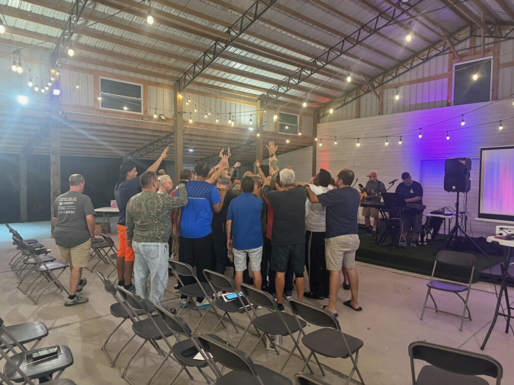 A group of men praying together with raised hands in a pavilion at Camp Impact
